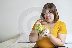 Obesity women eat green organic vegetables while sitting on the white bed. Fat female holding bowl salad vegetarian while