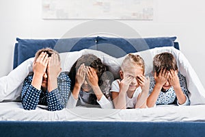 obscured view of multicultural children covering eyes while lying in bed under blanket photo