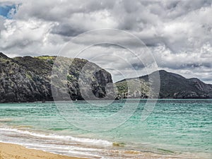 An ocean beach with rock cliffs, green hills abd cloudy sky