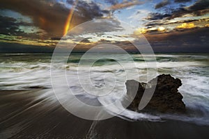 Ocean beach with waves, rock, and rainbow after storm