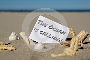 THE OCEAN IS CALLING text on paper greeting card on background of starfish seashell summer vacation decor. Sandy beach