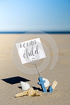 OCEAN CHILD text on paper greeting card in anchor paper holder and starfish seashell summer vacation decor. Sandy beach