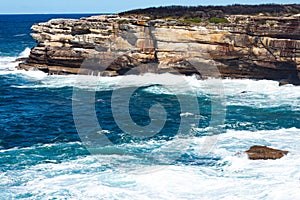 Oceanside rocky sandstone cliff with blue sea water waves creating whitewash against coastline and clear sky in background