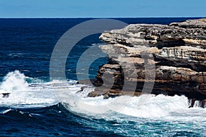 Oceanside rocky sandstone cliff with blue sea water waves creating whitewash against coastline and clear sky in background