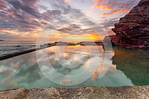 Ocean rock pool built into cliffs with small cave and sunrise reflections