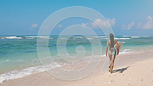 Ocean surf. Beautiful young surfer girl running across the blue sea beach with surfboard to the water. Sun shining