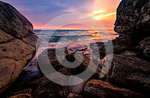 Ocean water splash on rock beach with pink and golden sunset sky. Sea wave splashing on stone at sea shore on summer. Tires and