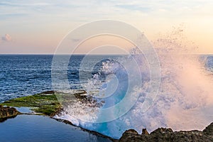 Ocean wave breaking on shorline cliff. Blue-green water, white spray in air. Pool of water in foreground. Ocean, sky in background