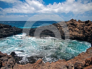 Ocean waves breaking on the rocky coast of hardened lava with caverns and cavities. Blue sky with white clouds and mountains