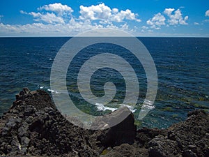 Ocean waves breaking on the rocky coast of hardened lava with caverns and cavities. Deep blue sky with white clouds on the horizon