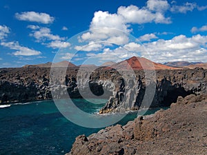 Ocean waves breaking on the rocky coast of hardened lava with caverns and cavities. Deep blue sky with white clouds and mountains