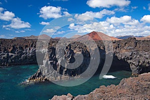 Ocean waves breaking on the rocky coast of hardened lava with caverns and cavities. Deep blue sky with white clouds and mountains