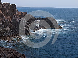 Ocean waves breaking on the rocky coast of hardened lava with caverns and cavities. Mountains and volcanoes on the horizon