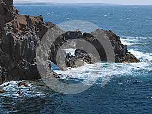 Ocean waves breaking on the rocky coast of hardened lava with caverns and cavities. Mountains and volcanoes on the horizon