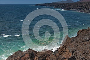 Ocean waves breaking on the rocky coast of hardened lava with caverns and cavities. Mountains and volcanoes on the horizon