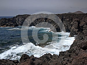 Ocean waves breaking on the rocky coast of hardened lava with caverns and cavities. Mountains and volcanoes on the horizon