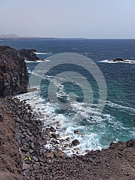 Ocean waves breaking on the rocky coast of hardened lava with caverns and cavities. Mountains and volcanoes on the horizon