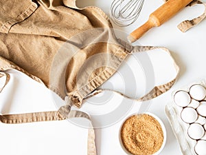 Ocher apron and various baking ingredients - eggs, sugar and kitchen utensils on white background. Top view