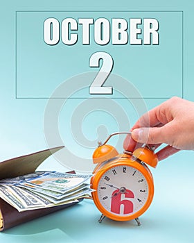 October 2nd. Hand holding an orange alarm clock, a wallet with cash and a calendar date. Day 2 of month.