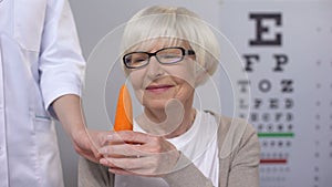 Oculist proposing carrot to smiling elderly lady, natural vitamins for sight