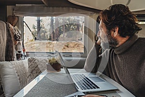 Off grid job lifestyle with mature man working on laptop inside a camper van and looking the forest outside the window. Van life