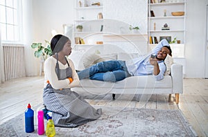 Offended black woman cleaning apartment while her lazy husband watching TV on sofa at home