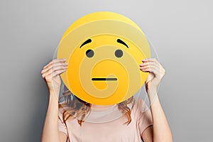 offended emoticon. Girl holds a yellow smiley with sad face  on a gray background