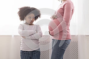 Offended little black daughter looking at pregnant mom`s belly with jealousy