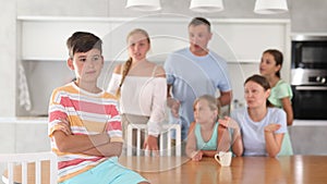 Offended pre-teen boy standing with arms crossed over chest at kitchen on background of his big family