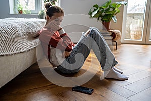 Offended unhappy teenage girl sadly looking at phone sits on floor after breaking up with boyfriend