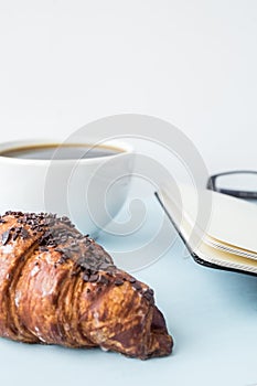 Office blue table with blank notepad, pencil, glasses, fresh croissant and coffee cup. Copy space for text.