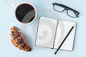 Office blue table with blank notepad, pencil, glasses, fresh croissant and coffee cup. Copy space for text. Flat lay