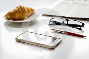 Office breakfast with croissant, phone and newspaper on white desk photo