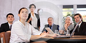 Office coworkers of different ages watching presentation while sitting at table with laptop and notes