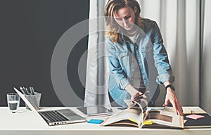 Office design studio, young businesswoman in denim shirt standing near desktop and leafing through catalog.