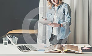 Office design studio, young businesswoman in denim shirt standing near desktop and leafing through catalog.