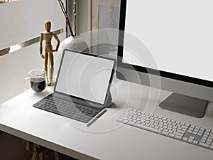 Office desk with blank screen tablet, computer device, coffee cup and decorations beside window