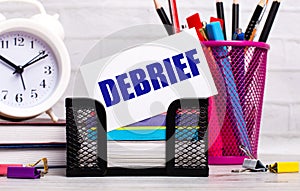 The office desk has diaries, an alarm clock, stationery, and a white card with the text DEBRIEF. Business concept