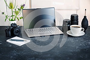 Office desk of a photographer with laptop, camera, lenses and a coffee cup on a dark table against a white wall, copy space,