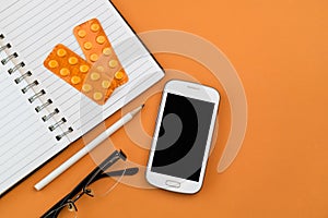 Office desk table with white smart phone,blank spiral notebook, black glasses ,white pencil and orange blisters