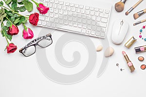 Office desk with wireless keyboard and mous, pink and red roses bouquet, women`s cosmetics on white background. Flat lay. Top vie