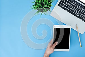 Flat lay top view mockup photo of working space with laptop and a hand holding on smart tablet on blue pastel background.