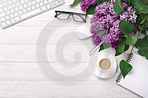 Office desktop with a bouquet of lilacs, coffee cup, keyboard, notebook and pen on white boards