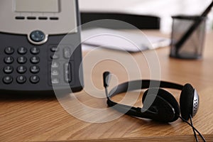 Office desktop telephone and headset on wooden table, closeup. Hotline service