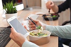 Office employee with smartphone having lunch at workplace, closeup.