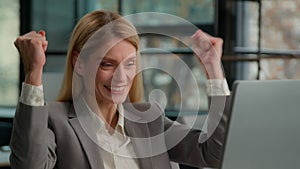Office female worker Caucasian middle-aged smiling businesswoman sitting at desk working on computer celebrate online