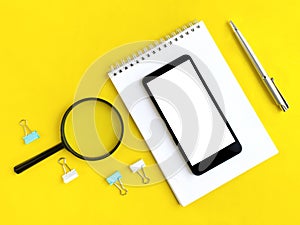 Office flat lay with smartphone, notepad, magnifier, pen and clips on yellow background.