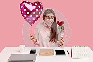 Office romance concept. Happy Caucasian lady holds valentine and bouquet of roses, recieves pleasant present from