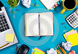 Office table desk with set of colorful supplies, white blank note pad, cup, pen, pc, crumpled paper, flower on blue
