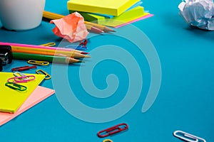 Office table desk with set of colorful supplies, white blank note pad, cup, pen, pc, crumpled paper, flower on blue